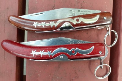The top is the original and the bottom is the knockoff. The top has a compressed wood handle, stained brown with rounded edges. The bottom is stained red with sharper angles. On the copy, the tang protrudes noticeably from the handle.