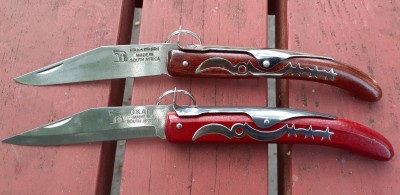 You can see a difference in the blades. The bottom has a sabre grind, compared the the top's flat grind. I'm pretty sure the bottom is a stainless steel, while all Okapis use 1055 carbon.